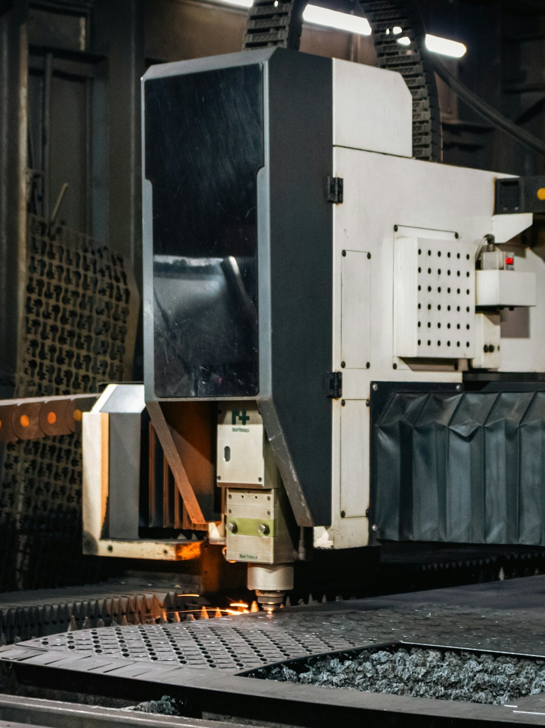 A close-up captures the action of an industrial CNC laser/plasma machine actively cutting a metal sheet. The cutting head is visible, emitting a small spark or glow of heat as it moves across the metal surface supported by a spiked cutting bed.